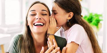 A woman laughs while a young girl whispers in her ear indoors.
