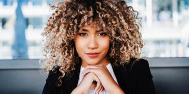 Woman with curly hair wearing a blazer, resting her chin on her hands and looking directly at the camera indoors.