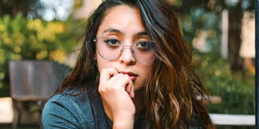 A woman with glasses looks thoughtfully toward the camera while resting her hand near her mouth outdoors.